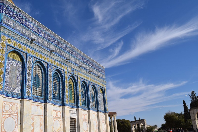 The Dome of the Rock stands against a bright blue sky. The sun emphasises the colours of the blue, white, green, and yellow tiles 
